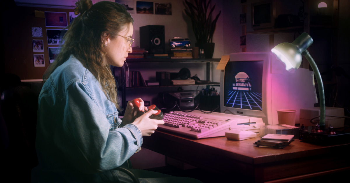 A cinematic, nostalgic photo of a person with a 90s-style hairstyle and denim jacket sitting at a wooden desk. They are using a vintage joystick in front of a classic beige CRT monitor displaying a retro-synthwave sunset graphic. The desk is cluttered with floppy disks, a thick mechanical keyboard, and a flexible desk lamp, creating a moody, authentic "early digital era" atmosphere.